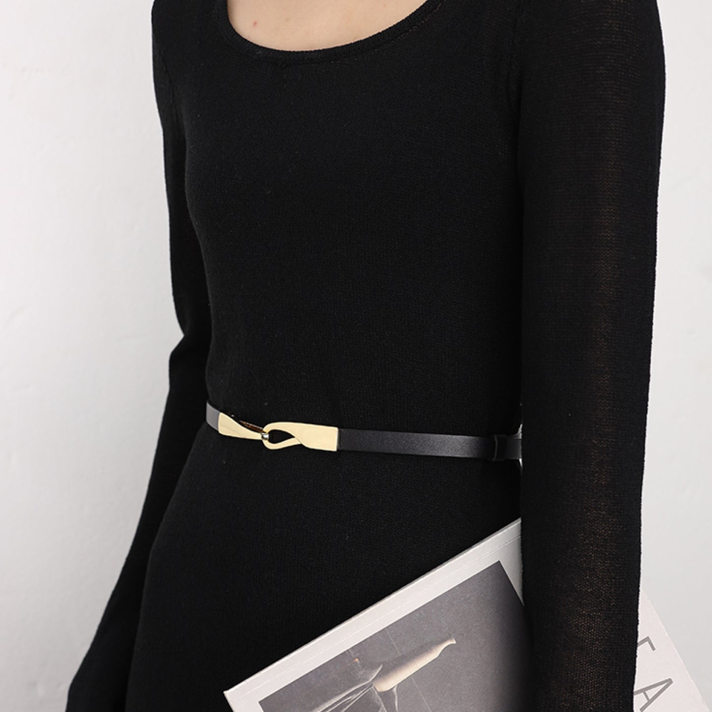 Person wearing a black long-sleeve top with a gold belt and holding a magazine.