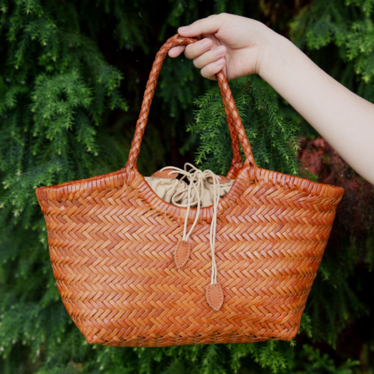 Hand holding a woven brown bag against a green leafy background