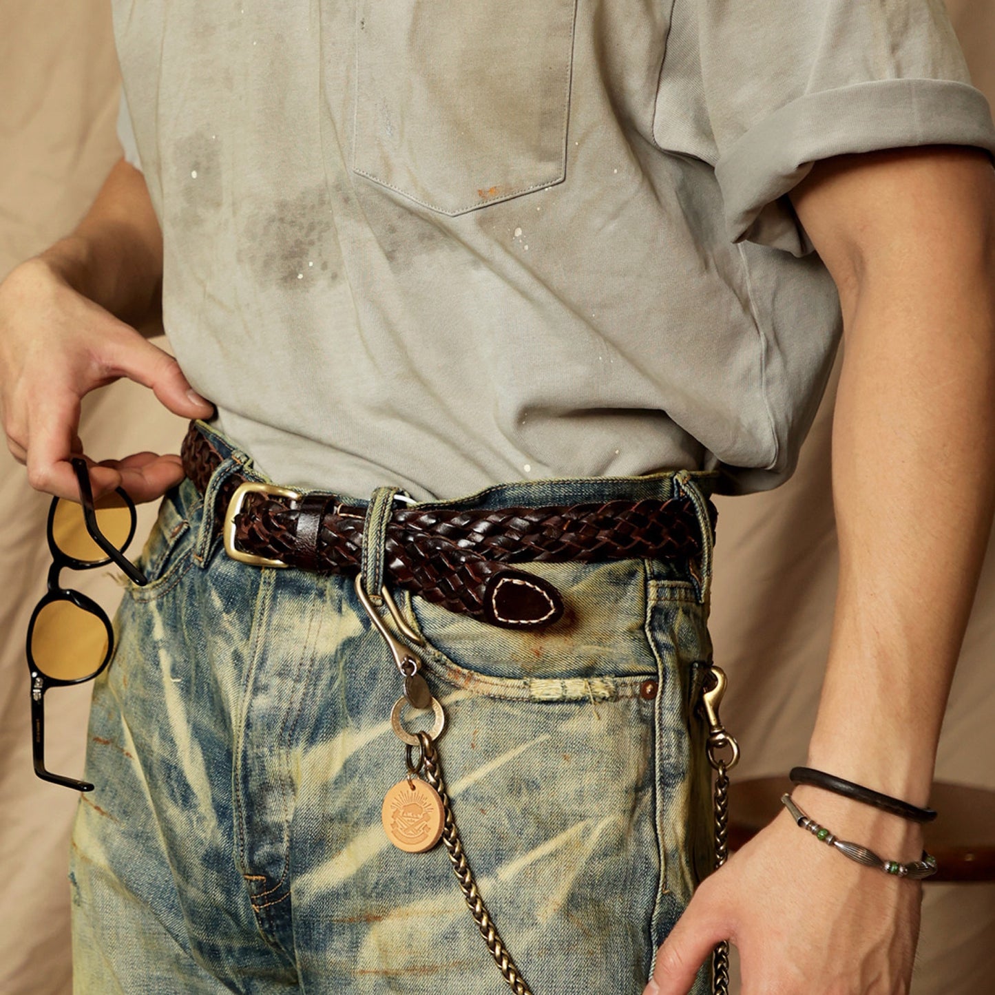 Person wearing a braided belt with a chain and round object, holding sunglasses.
