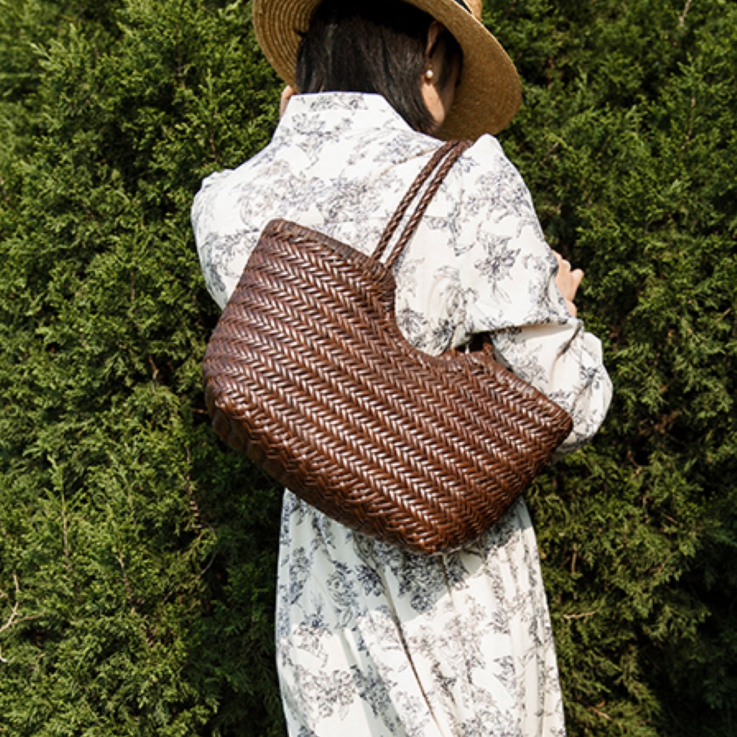 Person wearing a straw hat and holding a woven brown bag against a green hedge.