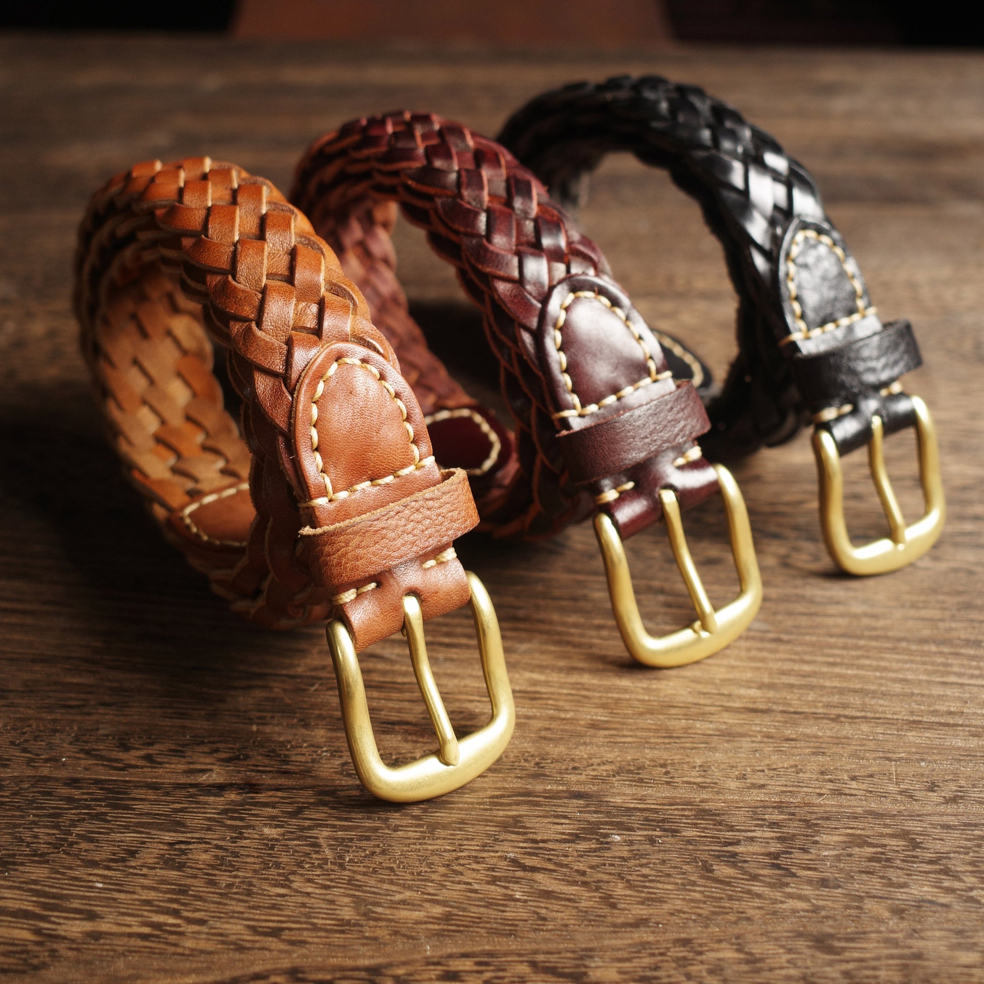 Three braided leather belts with gold buckles on a wooden surface