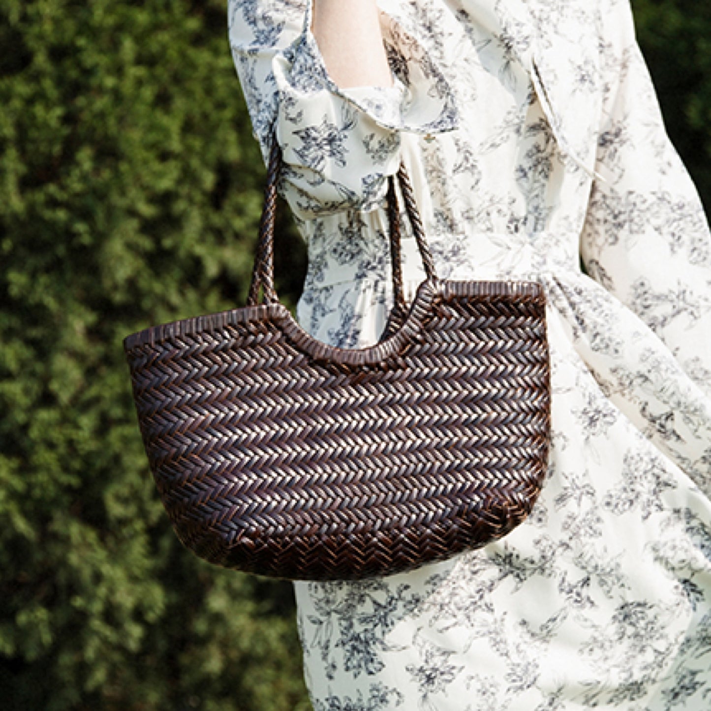 Woven handbag held by a person wearing a floral dress against a blurred green background