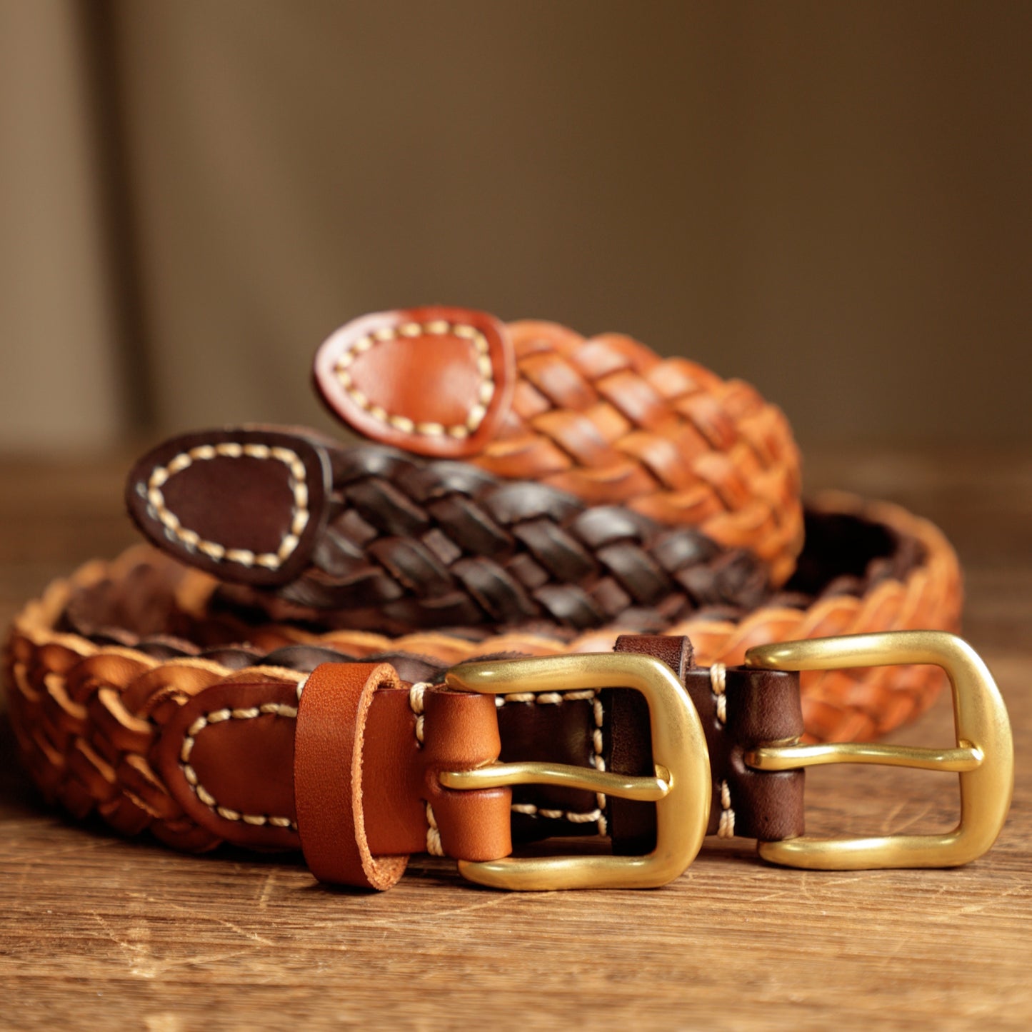 Two braided leather belts with gold buckles on a wooden surface.