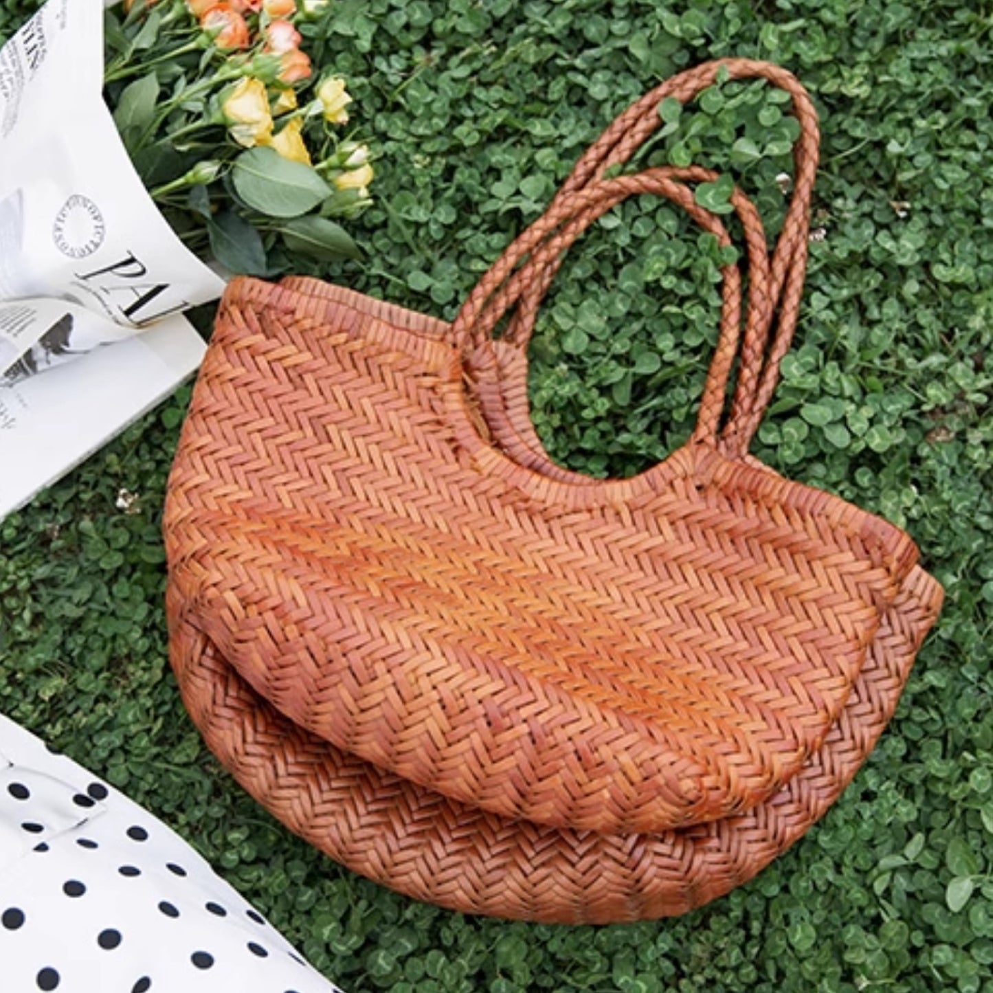 Woven brown bag on a grassy surface with flowers and a newspaper.