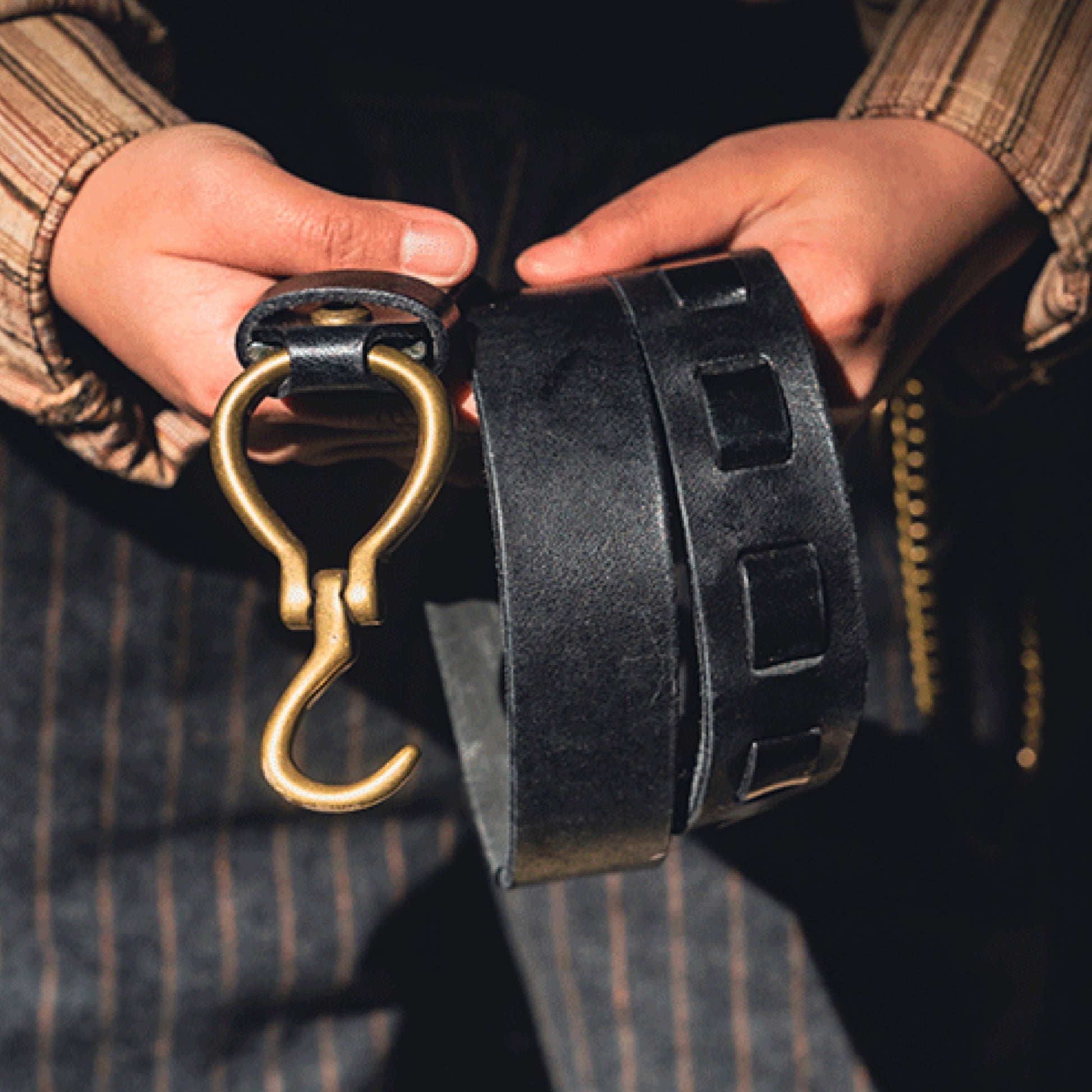 Black leather belt with a gold hook buckle held by a person wearing a striped shirt.